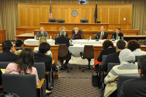 Ein Sitzungssaal mit einer Gruppe von Menschen, zwei ausländischen Flaggen, einem orangen Tisch, Wasserflaschen und einem Mikrofon auf dem Tisch.