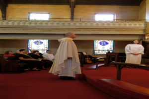 Eine Frau und ein Mann in weißen Kleidern stehen in einer Kirche, mit einem roten Teppich, Bänken, Musikinstrumenten und Fenstern im Hintergrund.