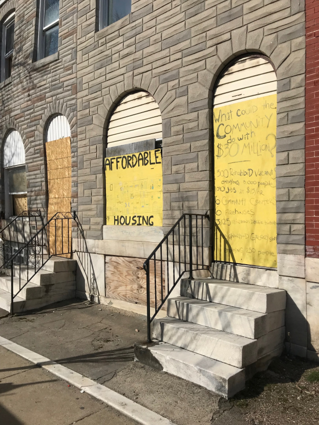 Ein zweistöckiges Gebäude mit vernagelten Fenstern, ein Schild mit der Aufschrift "Bezahlbarer Wohnraum" und eine Treppe, die zum Eingang führt, neben einer Straße.