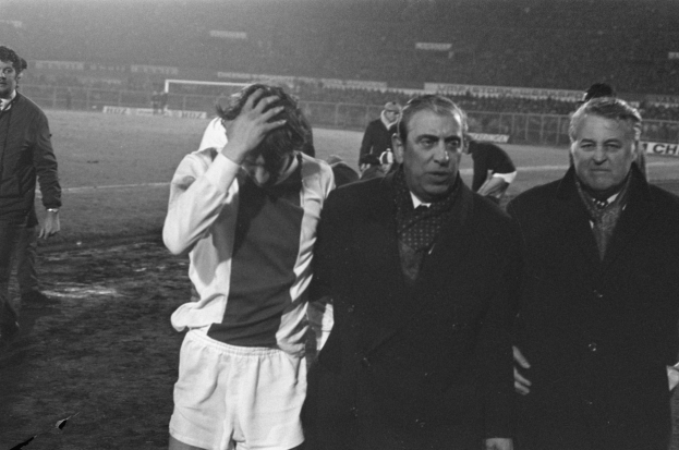 Schwarzes Bild einer Gruppe von Männern auf einem Fußballfeld, die nach einem Spiel enttäuscht wirken, mit einem Stadion im Hintergrund.