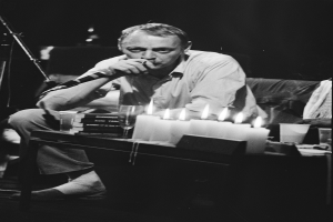 Ein Mann sitzt auf einem Sofa und hält ein Mikrofon in der Hand, singt, mit Kerzen, Gläsern und Büchern auf dem Tisch davor, dargestellt in Schwarz-Weiß.