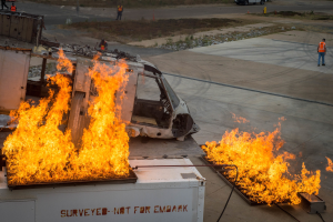 Ein Flugzeug in Flammen auf dem Rollfeld eines Flughafens, mit einem Fahrzeug und mehreren Personen in der Nähe, und Bäumen im Hintergrund.