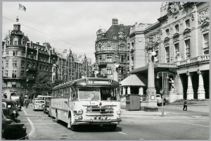Ein Schwarz-Weiß-Foto einer Stadtstraße mit einem Bus, der darauf fährt, umgeben von Gebäuden mit Fenstern, Bäumen, Laternen, einer Fahne mit einem Mast und ein paar Menschen.