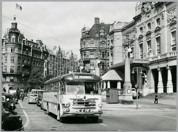 Ein Schwarz-Weiß-Foto einer Stadtstraße mit einem Bus, der darauf fährt, umgeben von Gebäuden mit Fenstern, Bäumen, Laternen, einer Fahne mit einem Mast und ein paar Menschen.