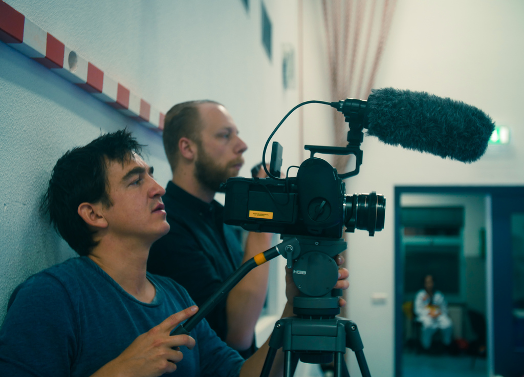 Ein Mann in einem blauen T-Shirt filmt mit einer Kamera, über ihm ist ein Mikrofon zu sehen, während ein Mann in einem schwarzen Hemd und eine Frau in einem weißen Kleid in der Nähe sind und eine weiße Wand mit Vorhängen im Hintergrund zu sehen ist.