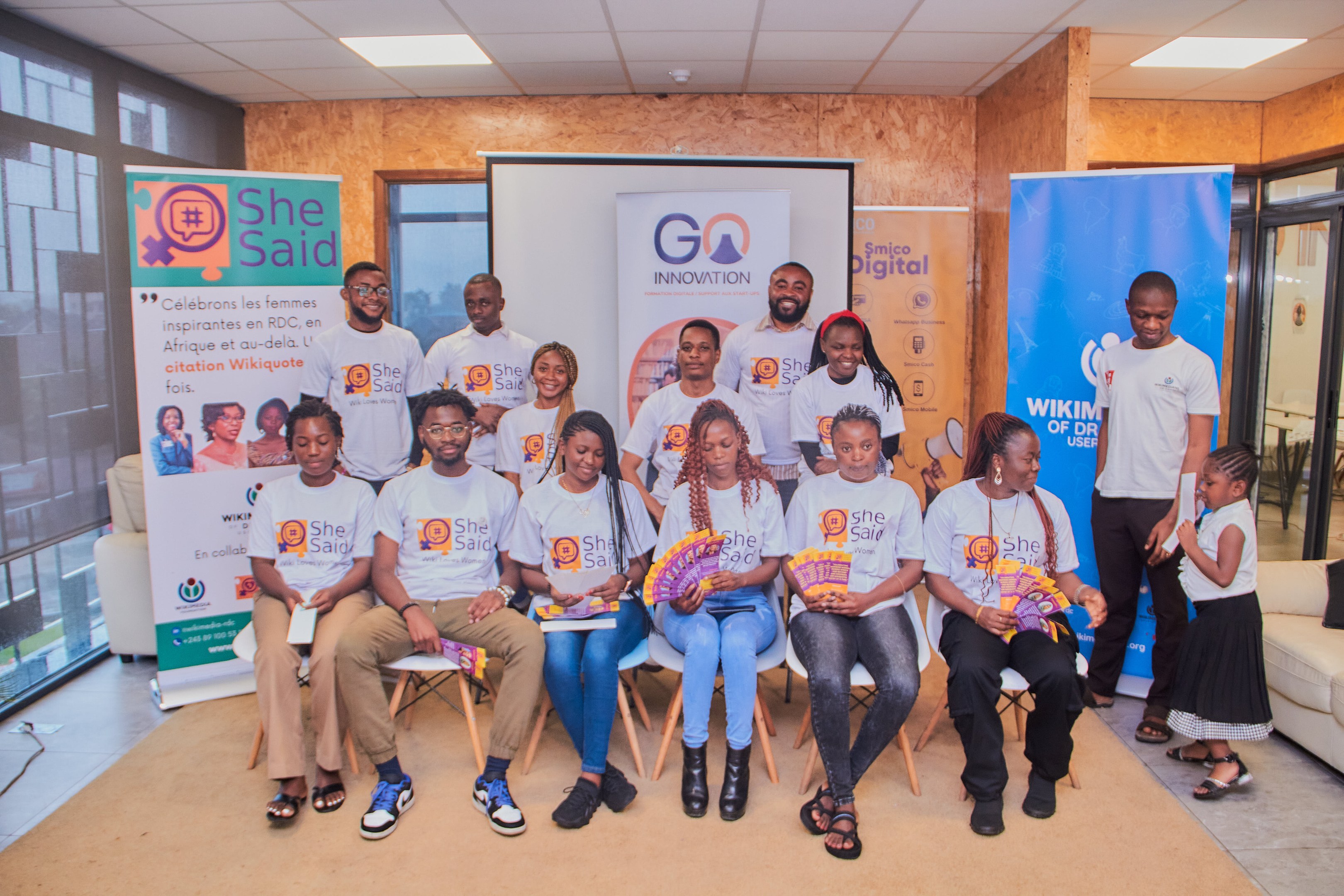 Gruppe von Menschen, die vor einem Bildschirm mit Schildern posieren, die "She Said - Go Innovation" lesen.