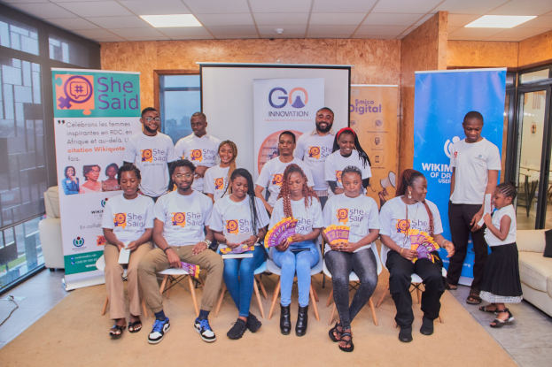 Gruppe von Menschen, die vor einem Bildschirm mit Schildern posieren, die "She Said - Go Innovation" lesen.