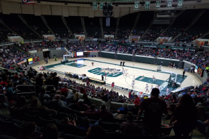 Eine große Arena voller Zuschauer, die ein Basketballspiel sehen, mit einem Spielfeld in der Mitte und Anzeigetafeln, Scheinwerfern und anderen Objekten oben.