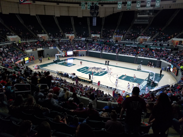 Eine große Arena voller Zuschauer, die ein Basketballspiel sehen, mit einem Spielfeld in der Mitte und Anzeigetafeln, Scheinwerfern und anderen Objekten oben.