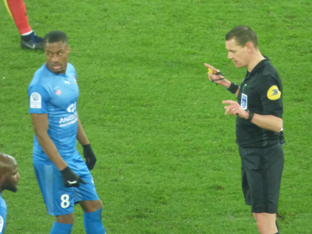 Ein Schiedsrichter spricht mit einem Fussballspieler auf dem Feld, umgeben von einer Gruppe von Menschen, wobei eine Person ein Objekt hält und die Beine einer anderen Person im Hintergrund zu sehen sind.