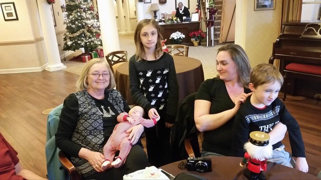 Ein Raum mit einer Wand, die ein Fotoframe, einen Weihnachtsbaum, eine Lampe und Blumenpflanzen zeigt. Im Vordergrund gibt es Treppen, ein paar Menschen, die an Tischen sitzen, mit einem Spielzeug und einer Kamera auf einem der Tische.