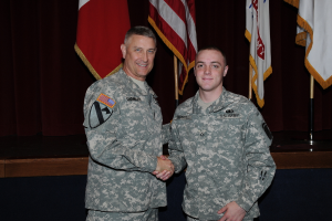 Zwei Personen in Uniformen geben sich die Hand und lächeln, vor vier Fahnen auf Stangen gegen einen dunklen Hintergrund.