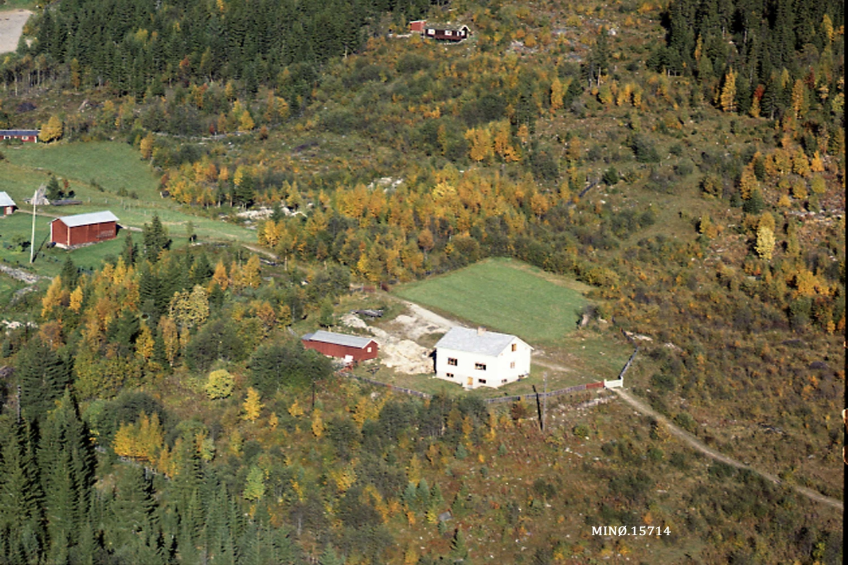 Eine Luftaufnahme eines kleinen Bauernhofs, umgeben von Bäumen, Gras und Häusern, mit Text am unteren Bildrand.