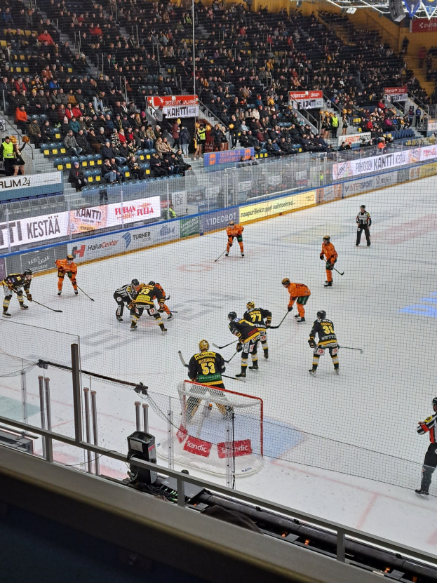 Eine Gruppe von Menschen, die Hockey auf einem Eisring spielen, umgeben von einem Zaun, mit Zuschauern im Hintergrund und beleuchtet von Stadionlampen.