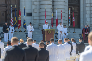 Gruppe von Männern in weißen Uniformen vor einem Gebäude mit Säulen und Türen, ein Podium mit Mikrofon in der Mitte, Fahnen mit Stangen im Hintergrund, Stühle und Treppen zum Gebäude hin.