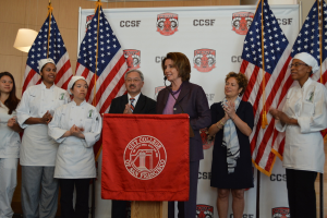 Eine Frau steht an einem Rednerpult mit einem Mikrofon, lächelt, vor einem roten Tuch, Flaggen, einem Banner, einer Lampe und einer Wand im Hintergrund.