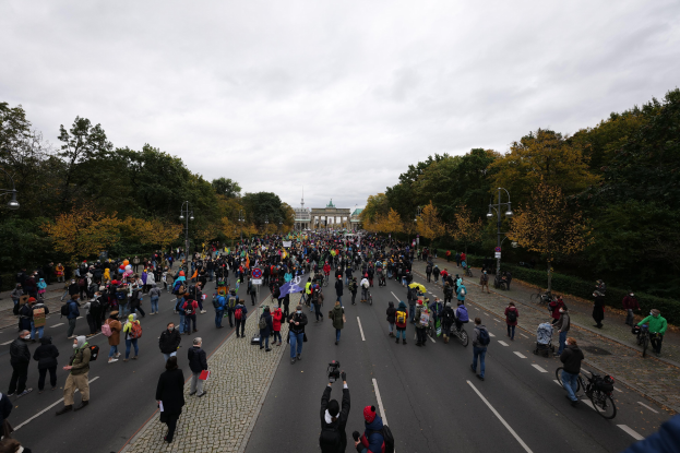 Eine große Gruppe von Menschen marschiert auf einer von Bäumen gesäumten Straße mit Laternenmasten, wobei einige Kameras halten, während einer Protestmarsch in Berlin mit einem Gebäude und einem klaren Himmel im Hintergrund.