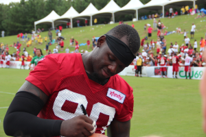 Ein Mann in einem roten Hemd unterschreibt einen Fußball auf einem Feld umgeben von Menschen, Bannern, Zelten, Bäumen und einem klaren blauen Himmel.