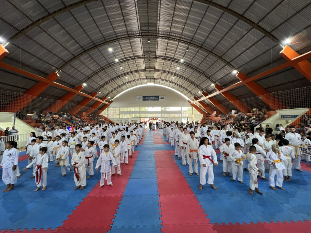 Eine gro├če Gruppe von Menschen, die auf einer blauen und roten Matte in einer Turnhalle mit Zuschauern auf Treppen und Deckenleuchten steht, bei einem Taekwondo-Event, das durch Schilder im Hintergrund angezeigt wird.