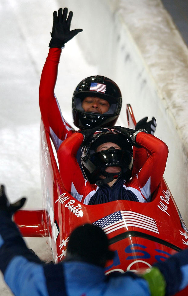 Zwei Bobsleighs fahren eine Bahn hinunter, einer mit sichtbarer Helm und Handschuhen, eine Person unten und eine Wand im Hintergrund.