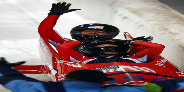 Zwei Bobsleighs fahren eine Bahn hinunter, einer mit sichtbarer Helm und Handschuhen, eine Person unten und eine Wand im Hintergrund.