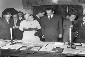 Schwarz-weiß-Fotografie einer Registratur-Szene mit Menschen, die Dokumente an einem Tisch mit Büchern, Kerzenständern und anderen Gegenständen unterzeichnen, mit einer Wandinschrift im Hintergrund.