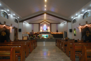 Das Innere einer Kirche mit hölzernen Kirchenbänken, Blumensträußen, Lampen, Lautsprechern und gerahmten Gegenständen an den Wänden, unter einer Decke mit Lampen.