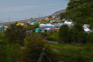 Ein Blick auf Häuser, eine Straße, Pfähle, einen Zaun, Bäume, Gras und einen Gewässerabschnitt.