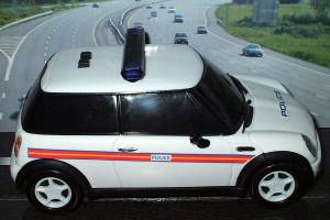 Ein weißer Polizeiwagen steht im Vordergrund mit vielen Autos auf der Straße dahinter, umgeben von Gras und Bäumen auf beiden Seiten, in einer ländlichen Umgebung.