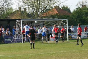 Spieler spielen Füßball auf einem Feld mit einem Tor, während Zuschauer dahinter stehen, mit Bäumen und Häusern im Hintergrund.