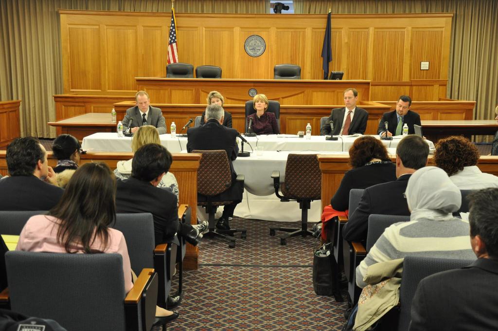 Ein Versammlungsraum mit einer Gruppe von Menschen, die auf Stühlen sitzen, zwei ausländischen Flaggen, einem orangen Schreibtisch, Wasserflaschen und einem Mikrofon auf dem Schreibtisch.