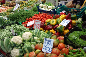 Ein belebter Bauernmarkt mit verschiedenen Gemüsen wie Tomaten, Paprika, Blumenkohl, und grünen Bohnen in Körben, Preisetiketten sichtbar, und einige Menschen in der Nähe.