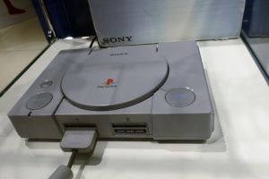 Eine graue PlayStation-Konsole auf einem weißen Tisch mit einem angeschlossenen Kabel und einigen undeutlichen Gegenständen im Hintergrund.