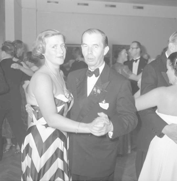 Ein Mann in einem schwarzen Blazer, weißem Hemd und schwarzen Hosen tanzt mit einer Frau in einem unbekannten Outfit auf einer formellen Veranstaltung, indem sie sich an den Händen halten und zur Musik an einer Wand schwenken.