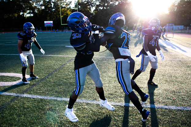Junge Männer in Helmen und Fußballschuhen spielen ein Spiel auf einem Rasenfeld mit Bäumen und einem klaren blauen Himmel im Hintergrund.