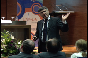 Ein Mann in einem schwarzen Blazer steht an einem Podium mit Mikrofonen, spricht in ein tragbares Mikrofon, mit einer Blumenanordnung und einem Tisch mit einem Buch und einer Wasserflasche in der Nähe, während drei Personen auf Stühlen vor ihm sitzen, vor einem Hintergrund aus einer braunen Wand und einer bunten Tafel mit Text.