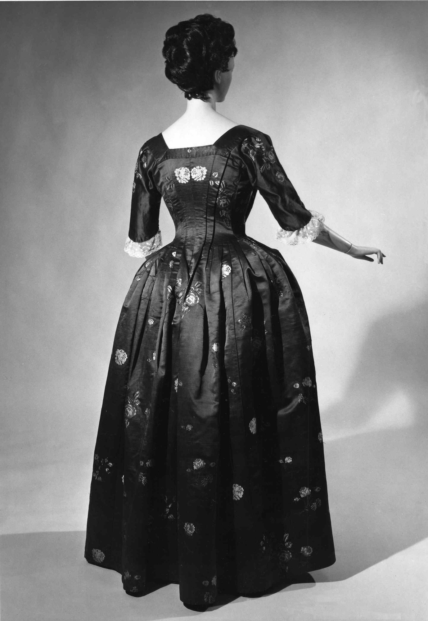 Eine Frau in einem Schwarz-Weiß-Foto aus den 1950er Jahren trägt ein langes, enges Kleid mit einem weiten Rock, hält ihre Hände zusammen und hat ihr Haar in einem klassischen Dutt mit dezentem Make-up und schwarzen Pumps.