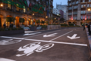Eine Stadtstraße mit einem Fahrradweg, Gebäuden, Straßenlaternen, Verkehrsschildern, Menschen auf dem Gehweg, Bänken, Pflanzen, Bäumen, dekorativen Elementen und einem klaren blauen Himmel.