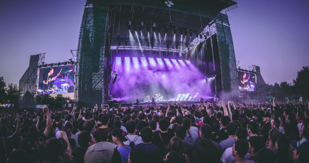 Eine große Menschenmenge auf einem Musikfestival schaut auf einer hell erleuchteten Bühne mit Instrumenten und Bildschirmen, die vor einem Gebäude, Bäumen und einem blauen Himmel aufgebaut sind.
