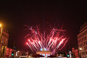 Eine belebte Stadtstraße in Berlin an Silvester, voller Menschen, Fahrzeuge und Gebäude, beleuchtet von Feuerwerk und Gebäudelichtern, die eine festliche Atmosphäre schaffen.