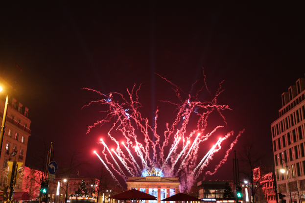 Eine belebte Stadtstraße in Berlin an Silvester, voller Menschen, Fahrzeuge und Gebäude, beleuchtet von Feuerwerk und Gebäudelichtern, die eine festliche Atmosphäre schaffen.
