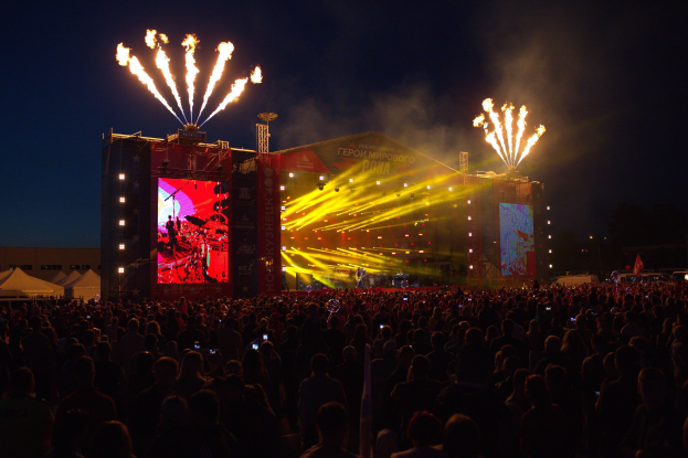 Große Menschenmenge auf einem Musikfestival mit beleuchteter Bühne und Performern, Bannern, Zelten links und Feuerwerk am dunklen Himmel.