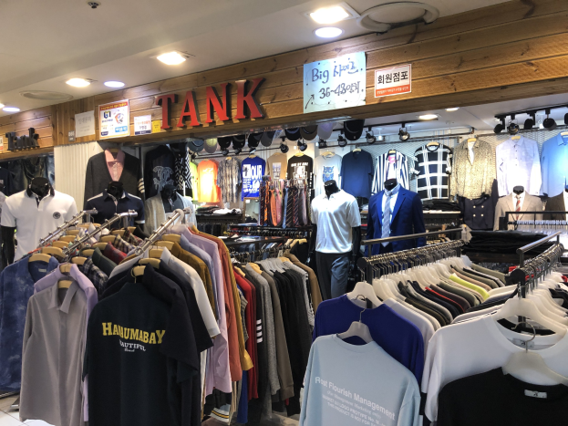 Ein moderner Bekleidungsladen mit zahlreichen Shirts auf Gestellen und Schaufensterpuppen, die die neuesten Modetrends präsentieren, mit Texttafeln an den Wänden und Deckenleuchten.