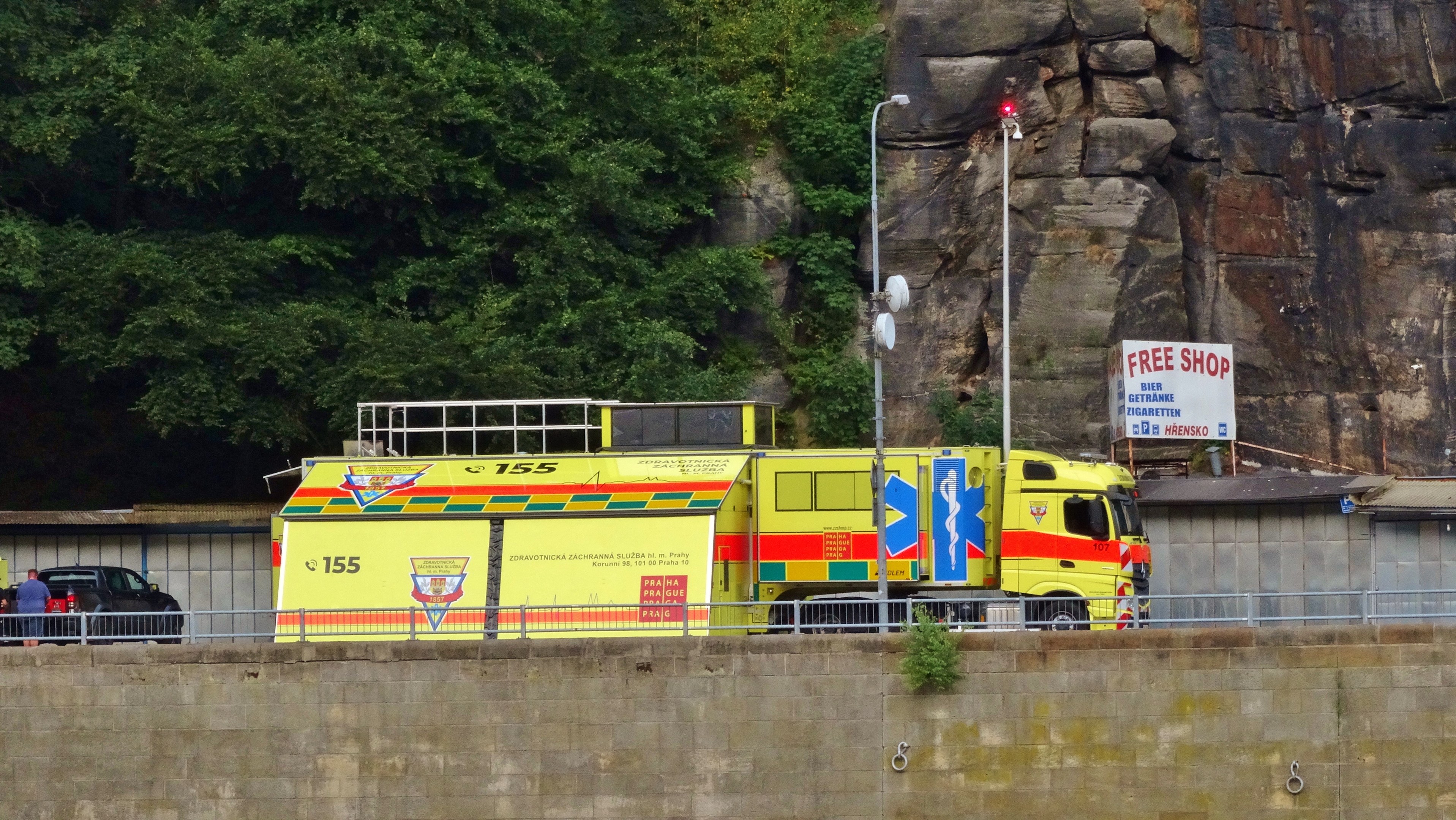 Ambulanz auf der Seite einer Straße neben einer Klippe geparkt, mit verschiedenen Gegenständen wie einer Wand, einem Geländer, Pfosten, Lampen und Schildern im Vordergrund und Bäumen und einem Auto im Hintergrund.