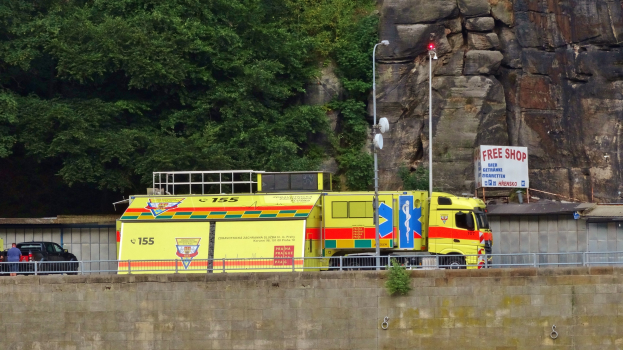 Ambulanz auf der Seite einer Straße neben einer Klippe geparkt, mit verschiedenen Gegenständen wie einer Wand, einem Geländer, Pfosten, Lampen und Schildern im Vordergrund und Bäumen und einem Auto im Hintergrund.