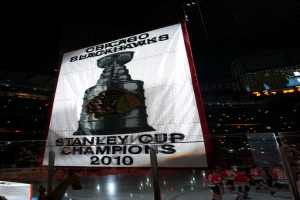 Eine nächtliche Stadion-Szene mit einem großen Banner in der Mitte, Zuschauern im Hintergrund, einem Glas auf dem Boden, einigen Spielern auf dem Feld und beleuchteten Tribünen.