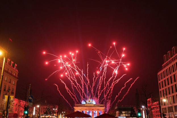 Eine belebte Stadtstraße bei Nacht am Silvesterabend in Berlin, mit Gebäuden, Bäumen, Laternen, Ampeln, Schildern, Zelten, Menschen und einem prächtigen Feuerwerk, das den Himmel erhellt.