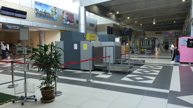 Leerer Flughafen-Terminal mit Check-in-Schalter, Topfpflanzen, Absperrpoller mit Bändern, stehenden Menschen, Gepäckwagen, Stühlen, Informationsanzeigen an den Wänden und Deckenbeleuchtung.