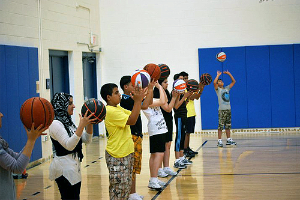 Eine Gruppe junger Menschen steht auf einem Basketballfeld, jeder hält einen Basketball in der Hand, im Hintergrund sind Türen und eine Wand zu sehen.