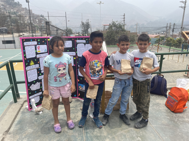 Eine Gruppe von Kindern mit Papierbeuteln auf einem Basketballfeld mit Beuteln und Gegenständen rechts und einer Tafel mit Text links, mit Zaun, Pfosten, Straßenlaternen, Bäumen, Gebäuden, Hügeln und Himmel im Hintergrund.